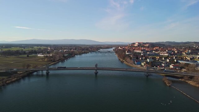 Ptuj, Slovenia, features its historic castle and old town with red rooftops, bridges over the Drava River, and surrounding hills and lake, captured in a daytime aerial cityscape from a drone.