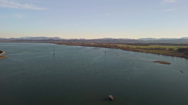 Ptujsko Jezero near Ptuj, Slovenia, features calm freshwater bordered by fields, wetlands, and distant hills under a clear blue sky in this wide aerial panorama captured by drone.