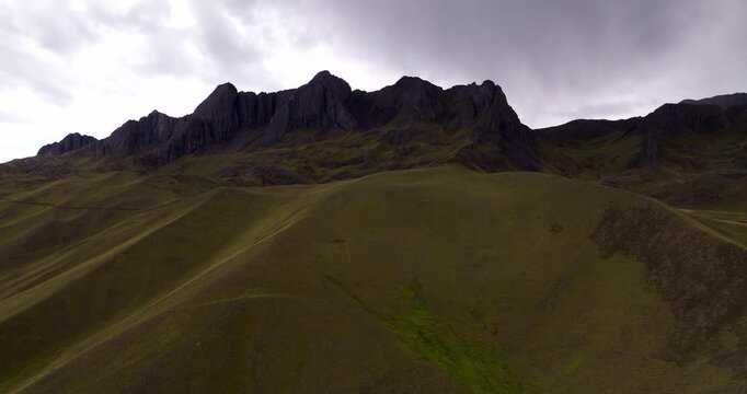 Cinematic aerial shot in Langui District, Peru, revealing rugged green-brown mountain slopes and jagged highland peaks beneath a moody overcast sky