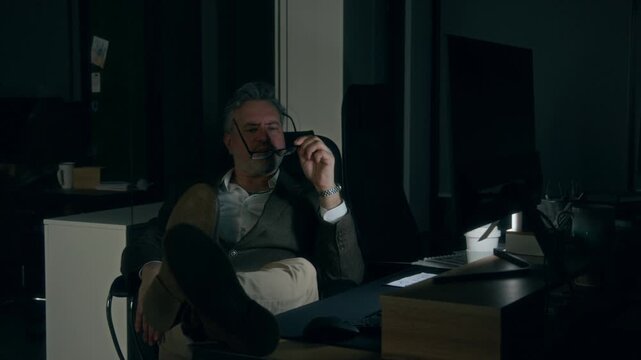 Full shot of frustrated 50-year-old Caucasian male manager sitting in dark office with feet on desk, staring at computer screen, pondering, encountering work problem or challenge, taking off glasses