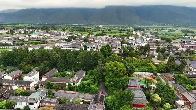 A highangle drone shot showcasing the traditional architecture of Baisha Ancient Town with Fuguo Monastery nestled among lush green trees against a mountain backdrop.