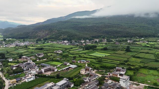 High angle aerial shot of traditional Chinese village architecture and agricultural fields nestled below misty mountains in Lijiang, Yunnan.