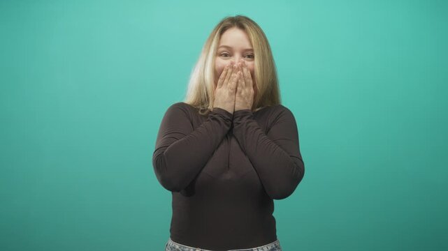 Woman covers mouth with hands in turquoise studio, smiling with eyes and visible laughter while standing in casual denim jeans, frontal pose; surprise joy.