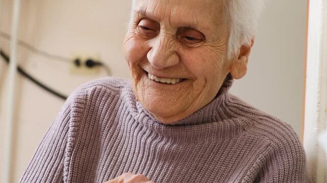Senior woman with wrinkled skin and gray hair sitting at table smiling happily and sincerely surrounded by love and care. Her eyes shine with kindness and joy. Care and love for lonely elderly