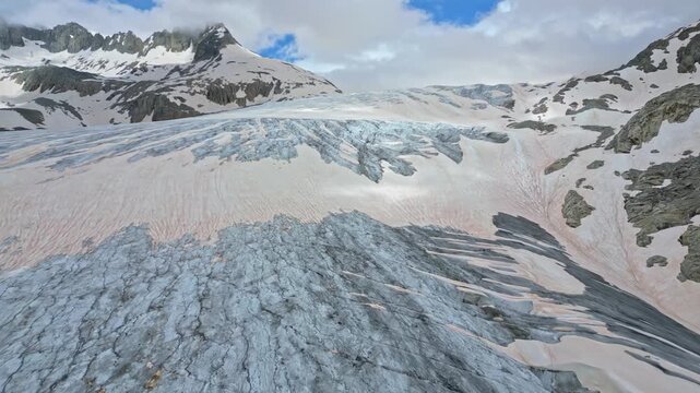 Aerial view of a vast glacier flowing amidst snow-capped mountains, its icy surface contrasting with the rugged peaks, Obergoms, Valais, Switzerland.