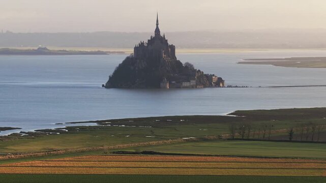 Aerial view of the historic Mont Saint-Michel abbey rising above the tidal waters and surrounding salt marshes during a hazy afternoon in Mont Saint-Michel, Normandy, France.