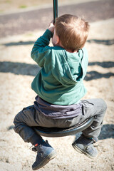 Fototapeta premium A young child swings on a round swing at a playground. The child grips the swing while enjoying the activity. Other children play in the background as the sun shines.