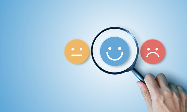 Hand holding magnifying glass focusing on happy smile face over neutral and sad icons on blue background. Concept of customer satisfaction survey, finding positive feedback and excellent service.
