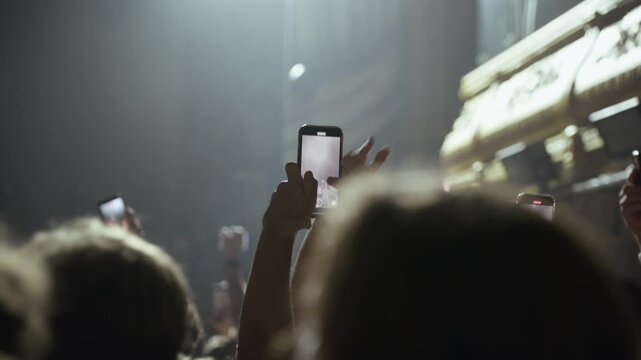 Crowd at a live concert or event captures the performance on their smartphones. Hands hold up phones, recording the stage illuminated by dynamic spotlights and smoke