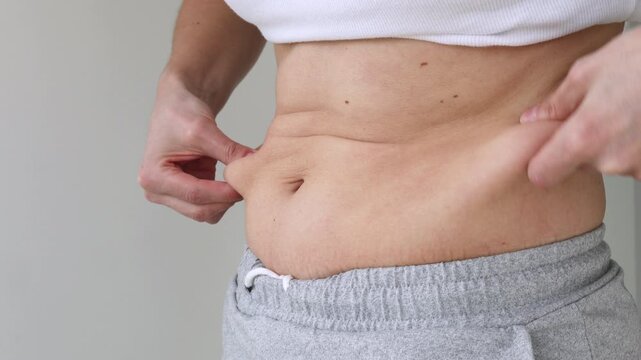 Close-Up of Woman's Hands Pinching Belly Fat Skin Fold at Waist in White Crop Top and Gray skirt &mdash; Female Checking Abdominal Subcutaneous Fat, Body Weight Awareness, Healthy Lifestyle Motivation