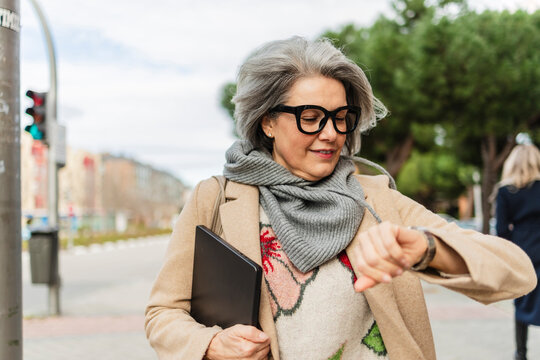 Freelancer in coat with laptop checking time on urban street