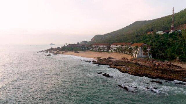 Aerial view of the coastal resort, with its sandy beach meeting the deep blue sea, nestled against a backdrop of lush green hills, Bai Xep Beach, Binh Dinh, Vietnam.