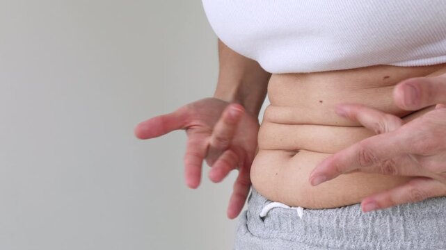 Close-Up of Woman's Hands Pinching Belly Fat Skin Fold at Waist in White Crop Top and Gray skirt &mdash; Female Checking Abdominal Subcutaneous Fat, Body Weight Awareness, Healthy Lifestyle Motivation