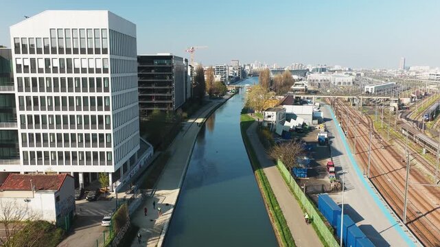 Aerial view of Canal de l'Ourcq, a tranquil waterway flanked by modern buildings and green spaces, reflecting the clear sky, Bobigny, Ile-de-France, France.