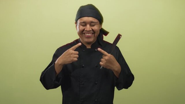 Young hispanic man chef in black uniform pointing to his smile while holding a cleaver in studio; pride craft.