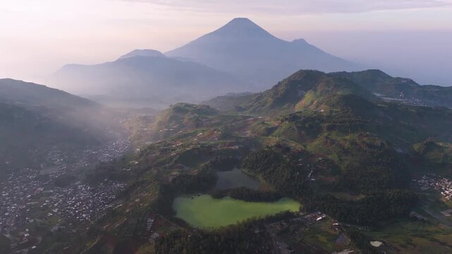 Aerial view of a green lake nestled among rolling hills and distant volcanoes, showcasing the natural beauty of Dieng, Central Java, Indonesia.