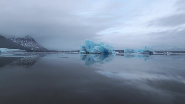 Aerial view of blue icebergs reflecting in the calm water of the Jokulsarlon glacial lagoon with mountains under an overcast sky in Jokulsarlon, Sveitarfelagid Hornafjordur, Iceland.
