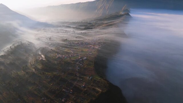 Aerial view of a village nestled in the mountains, shrouded by mist and fog, with the sun casting long shadows across the landscape, Bromo Tengger Semeru, Indonesia.