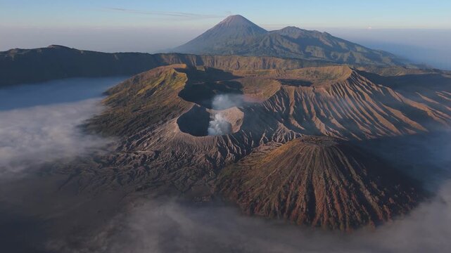 Aerial view of the volcano with a plume of smoke rising from its crater, surrounded by mist and rugged landscape, Bromo Tengger Semeru National Park, Indonesia.