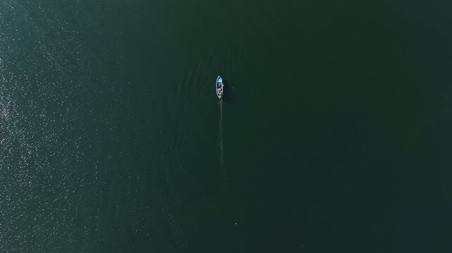 A small fishing boat travels through a large body of water. The sun is shining, and the water shows ripples around the boat. The scene is open with no other boats nearby.