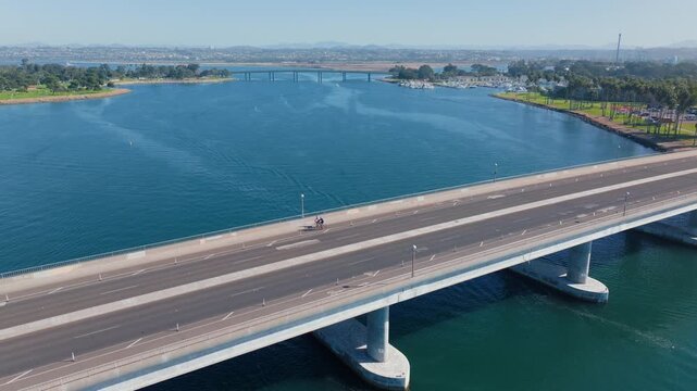 Drone footage capturing cyclists riding across concrete bridge over turquoise waters of Mission Bay in San Diego.