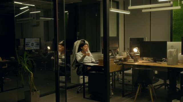 Wide shot of tired Hispanic female corporate manager falling asleep in chair in front of computer while working on project at night, waking, checking time on smartphone with shock and leaving office