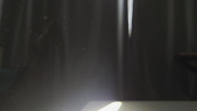 High-contrast, atmospheric footage of tiny dust particles illuminated by a single beam of light filtering through a window. The scene captures ethereal floating movement against a deep, shadowed backg