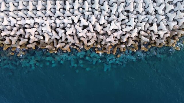 Cinematic top-down drone shot of a massive concrete breakwater at Agios Ioannis harbor. High-angle view of tetrapod blocks protecting the coastline from Aegean sea waves. Ideal for infrastructure.