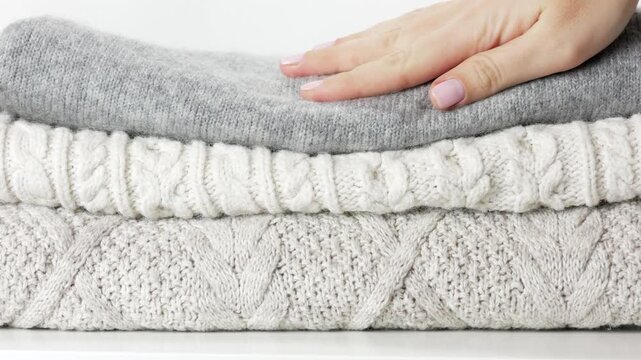 Woman hand touching pile of cozy sweaters in neutral tones. Cozy sweaters texture highlights soft wool fabric, seasonal fashion concept, comfort lifestyle and autumn winter clothing selection.