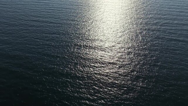 Sunlight shines down on the ocean, creating patterns on the water's surface. The scene shows a vast expanse with gentle waves moving under the bright light.