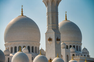 Sheikh Zayed Grand Mosque gleams under brilliant sunlight with white marble domes and minarets reflecting intense desert rays against clear blue skies © jovannig