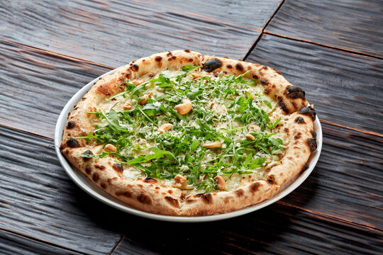 Top view of a crispy artisan white pizza topped with fresh arugula, roasted cashew nuts, and grated parmesan. Served on a white plate over a dark wooden background