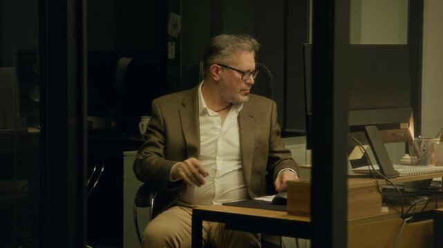 Medium shot of fatigued middle-aged Caucasian male financial analyst dozing off in front of computer at midnight while working on presentation, then waking, picking up document and leaving