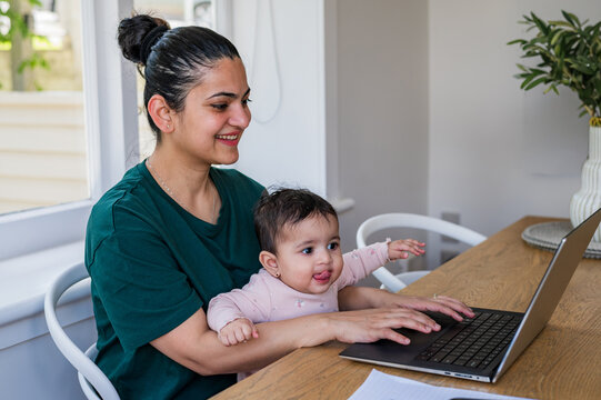 Indian mum working on her laptop on wooden table while holding baby girl on her lap