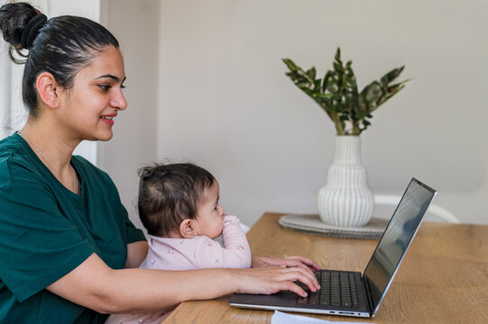 Indian mum working on her laptop on wooden table while holding baby girl on her lap