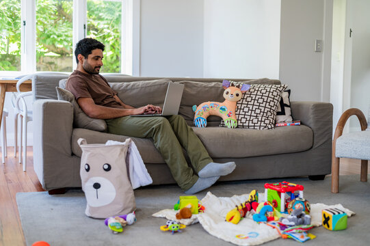 Indian man sitting on the couch in the living room working on his laptop with toys on the floor