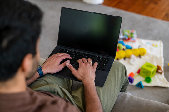 Indian man sitting on the couch in the living room working on his laptop with toys on the floor