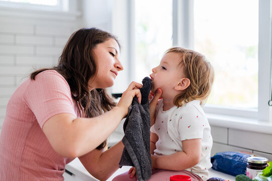 Mother wiping child&rsquo;s mouth with dark grey face towel on bright kitchen counter