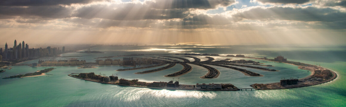 Panoramic helicopter view of Palm Jumeirah at sunset with dramatic sunlight breaking through clouds and urban skyline