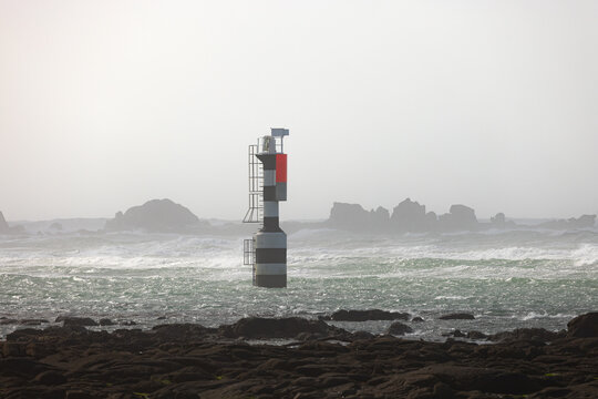 View of a black and white striped maritime beacon with a red top in rough sea with waves and rocky shore in Penmarch, Bretagne, France.