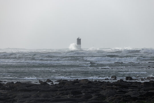 View of Phare d'Eckmuhl lighthouse with large waves crashing against it under a cloudy sky in Penmarch, Bretagne, France.