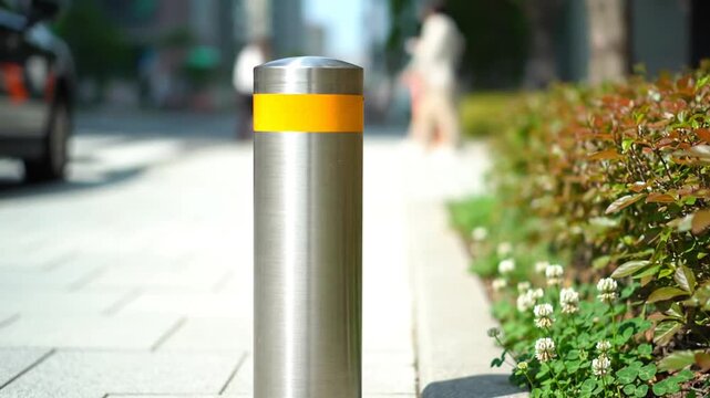 A silver bollard with a yellow stripe stands on a sidewalk next to a row of plants.