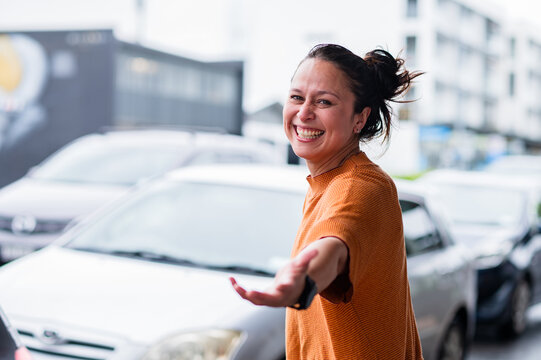 Woman standing in an urban street smiling and extending her hand towards the camera among cars