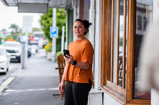 Woman standing on the street by a shop holding a mobile phone and looking ahead