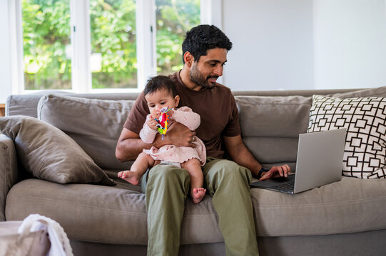 Man sitting on a couch with a baby in his lap while using a laptop in the living room