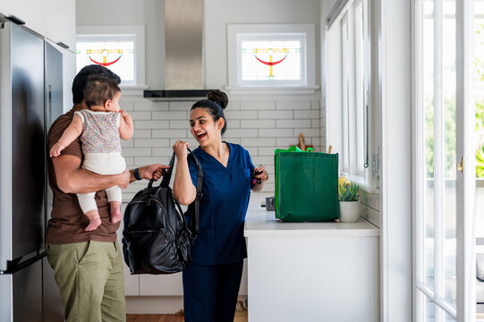 Person carrying a backpack and smiles at another who holds a baby in a kitchen setting
