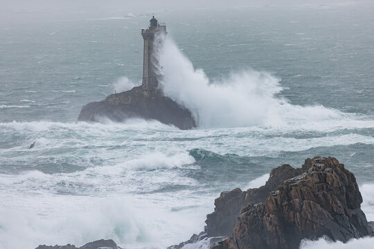 View of Phare de la Vieille lighthouse at Pointe du Raz with massive waves crashing against the stone tower during a storm in Plogoff, Brittany, France.