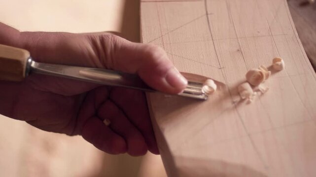 Woodworker's hand using a sharp metal tool to peel away thin shavings from a wooden block, focusing on texture and action. Slow motion close up.