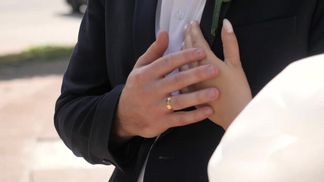 Elegant groom wearing dark tuxedo and gold wedding ring gently covers bride hand adjusting delicate white rose boutonniere on his lapel during romantic outdoor marriage ceremony.