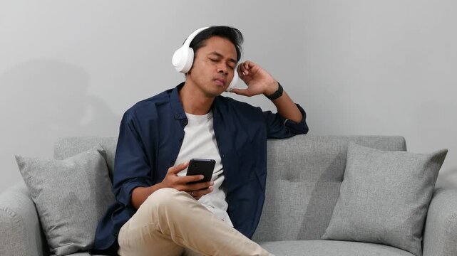 Happy carefree Young Asian man in stylish headphones listening to music from smartphone moving head